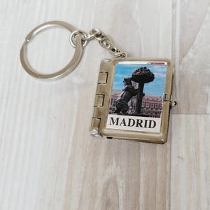 May include: Silver keychain with a rectangular charm depicting a bear and a tree, with the word "MADRID" beneath. The charm is connected to a chain and a circular keyring.