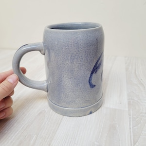 Puede incluir: Una jarra de gres azul grisáceo con un asa grande y un diseño pintado en azul oscuro. La jarra tiene un acabado moteado. Está sobre una superficie de madera clara.