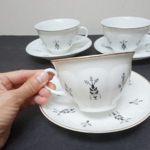 May include: Three white teacups and saucers with a black floral design. The cups have a gold rim and the saucers have a gold band around the edge.