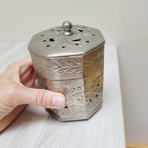 May include: A silver-colored, octagonal metal box with a lid. The box features decorative cut-out patterns and embossed floral designs. The lid has a small knob on top. The box is held by a hand.