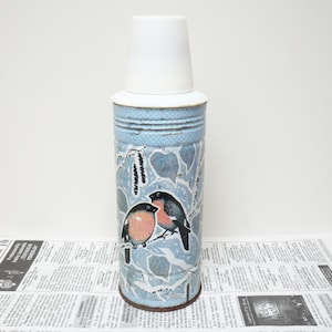 May include: A blue and white flask with a white lid and a design of two birds on a branch. The flask is sitting on a newspaper.