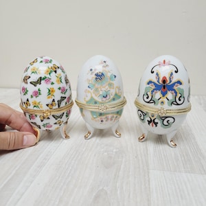 May include: Three decorative egg-shaped trinket boxes. The boxes are white with floral and butterfly designs. Each egg has a gold-colored metal hinge and three small gold-colored feet. The first egg has a butterfly and floral pattern, the second has a floral pattern, and the third has a floral design.