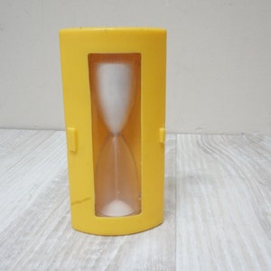 Op de afbeelding: Een gele zandloper met een doorzichtige, met zand gevulde glazen timer. De zandloper is ingekapseld in een rechthoekig geel frame met een gebogen basis. De timer is ongeveer 10 cm hoog.