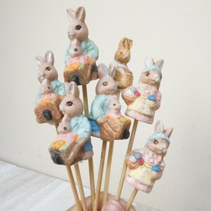 Könnte beinhalten: Ein Set aus acht Keramik-Hasenfiguren, jede mit einem Korb oder einer Schubkarre gefüllt mit bunten Ostereiern. Die Hasen sind in Pastellfarben bemalt, darunter Blau, Rosa und Gelb. Die Figuren sind an Holzspießen befestigt, was sie perfekt macht, um ein Frühlings-Mittelstück oder einen Ostertisch zu dekorieren.