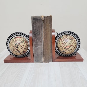 Può includere: Una coppia di fermalibri in legno con due piccoli mappamondi montati su basi rettangolari in legno marrone scuro. Ogni mappamondo è circondato da un anello nero con segni numerici da 0 a 90. Due libri dall'aspetto vissuto sono posti tra i fermalibri.