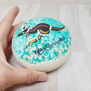 May include: A hand-painted, round stone featuring a kangaroo design. The kangaroo is depicted in black, brown, and yellow against a turquoise background with white speckles. The text "NGURRANGUNGA" is written in white.