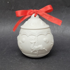 May include: White ceramic Christmas bauble featuring a raised design of a carousel with a reindeer. The bauble is tied with a red ribbon.