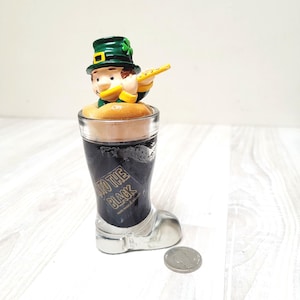 May include: A green leprechaun figurine playing a flute, sitting on top of a glass boot filled with dark liquid. The boot is labelled "Into the Black" and has a silver base. A silver coin is in the foreground.