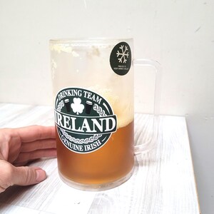 Może przedstawiać: Przezroczysty plastikowy kufel do piwa z zielono-białą naklejką z napisem "Drinking Team Ireland Genuine Irish". Kufel jest wypełniony jasnobrązowym piwem i ma naklejkę z płatkiem śniegu z napisem "Freezes to keep drinks cold".