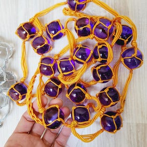 Può includere: Una lunga collana di perline di vetro viola, ciascuna racchiusa in una rete tessuta gialla, pende su una superficie bianca. Le perline sono infilate su un cordoncino giallo.