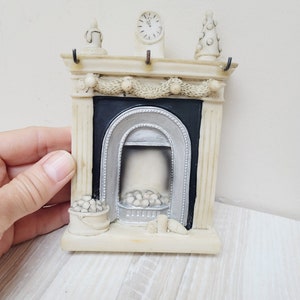 May include: A white ceramic fireplace mantel with a black and silver fireplace, a clock, and a garland of white beads. There are white balls in a bucket on the hearth.