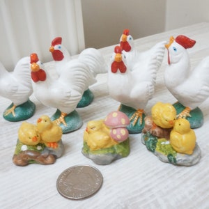 May include: A set of white ceramic rooster figurines with yellow chicks. The roosters have red combs and wattles. The chicks are sitting on green bases with flowers and mushrooms.
