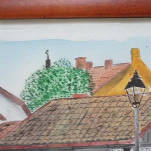 May include: Watercolor painting of a European town scene. The artwork depicts buildings with red tile roofs, a yellow building, and a street lamp. A green tree and a light blue sky are also visible.