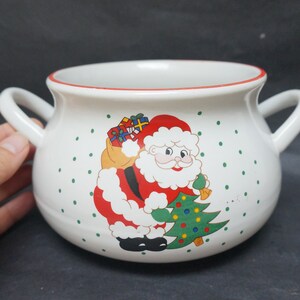 Könnte beinhalten: Weiße Suppenschüssel aus Keramik mit rotem Rand und einem weihnachtlichen Design mit dem Weihnachtsmann, der einen Sack mit Geschenken und einen Weihnachtsbaum trägt. Die Schüssel hat zwei Henkel.