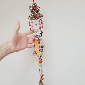 Puede incluir: Un móvil artesanal con una parte superior de metal calado, detalles de piedras rojas y cuentas coloridas. El móvil incluye una pluma, pequeñas campanas y varios elementos decorativos, creando una estética bohemia.