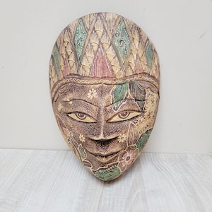 May include: A carved wooden wall mask featuring a brown face with almond-shaped eyes and a small mouth. The forehead is decorated with a diamond pattern in green and tan, while the cheeks and chin display intricate floral batik-style motifs.