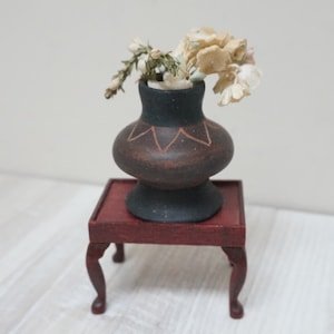 Puede incluir: Un pequeño jarrón de cerámica marrón oscuro con un diseño geométrico se encuentra sobre una mesa de madera roja. El jarrón está lleno de flores blancas secas.