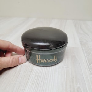 Op de afbeelding: Een kleine, ronde pot met een zwart deksel en een donkergroene body. De gouden tekst "Harrods" is op de zijkant van de pot gedrukt. De pot staat op een lichtgekleurd oppervlak.