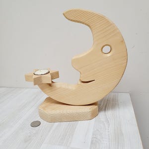 Könnte beinhalten: Kerzenhalter aus Holz in Form von Mond und Stern. Der Mond ist mit einem Gesicht geschnitzt und der Stern steht auf einem kleinen Sockel. Der Halter ist aus Naturholz.