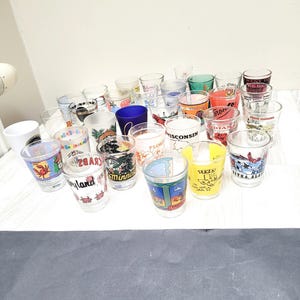 Könnte beinhalten: Eine Sammlung bunter Schnapsgläser mit verschiedenen Designs von Bundesstaaten und Städten. Die Gläser sind in einer Gruppe auf einer weißen Oberfläche angeordnet. Einige der Designs beinhalten Florida, Wisconsin, Maryland und Minnesota.