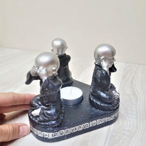 Può includere: Un portacandele nero e argento con tre statuette di monaci seduti. Ogni monaco ha la testa argentata e indossa una veste nera. Una piccola candela bianca è posta al centro del portacandele.