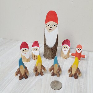Op de afbeelding: Een set houten dwergfiguren, waaronder een lange dwerg met een rode muts en een witte baard, en kleinere dwergen met rode, gele en blauwe mutsen. De dwergen zijn geschilderd met details zoals ogen, neus en mond.
