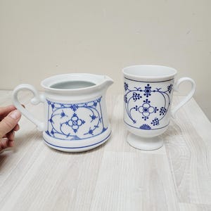 May include: A white ceramic creamer and mug set with blue floral patterns. The creamer has a handle and spout, while the mug features a pedestal base and a curved handle. The design is a classic, elegant style.