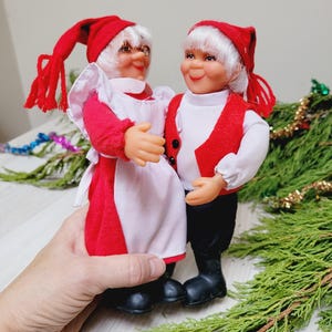 Op de afbeelding: Twee vintage kerstelfpoppen. De poppen zijn gekleed in rode en witte outfits met rode hoeden. De poppen houden elkaar vast. De poppen hebben wit haar en lachende gezichten. De poppen staan op een groene ondergrond.