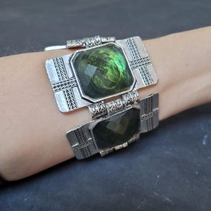 May include: Silver cuff bracelet with two rectangular, faceted, green stones. The bracelet has an ornate, textured design around the stones and on the band. The jewelry is worn on a wrist.