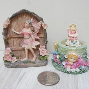 May include: Two fairy figurines, one standing in a doorway with pink flowers and the other sitting on a green base with pink flowers. Both figurines are made of resin and have painted details.
