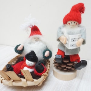 May include: Two knitted Christmas ornaments. A Santa Claus figure with a white beard and red hat sits in a woven basket with a snowman figure. A gnome figure with a red hat and gray beard holds a sheet of music and sits on a pine cone.