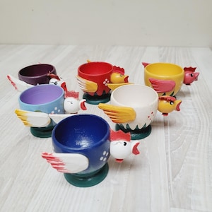Puede incluir: Colección de hueveras de madera pintadas a mano con forma de gallinas. Cada huevera está pintada de un color diferente, incluyendo rojo, azul, amarillo, morado y blanco, con alas y diseños de cabeza de gallina detallados. Las hueveras miden unos 8 cm de alto.