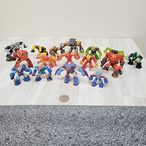 May include: A collection of colorful toy robots in various poses. The robots are primarily red, blue, yellow, green, and black, with detailed designs. They are arranged on a light-colored surface.