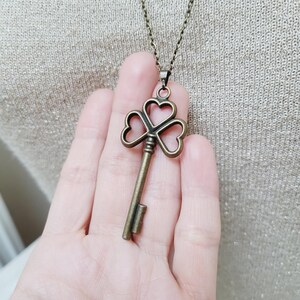 May include: A bronze key pendant with a clover-shaped top hangs from a delicate chain. The key's design features three heart-shaped cutouts. The necklace is displayed against a neutral background.