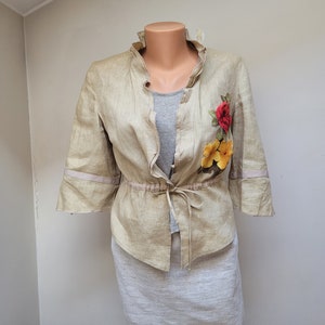 Può includere: Una giacca di lino beige con colletto arricciato e una cintura da annodare in vita. La giacca presenta un ricamo floreale sul lato destro con fiori rossi, gialli e arancioni.