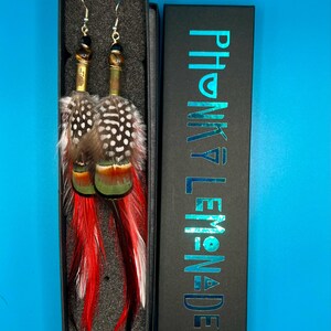May include: A pair of handmade feather earrings with red, white, and black speckled feathers. The earrings have a gold-colored metal base and are displayed in a black box with the text "PHONKY LEMONADE" in teal.