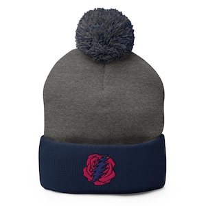 May include: A gray beanie with a navy blue brim and a dark blue pom-pom. The front of the beanie features an embroidered design of a red rose with a blue lightning bolt through it. This winter hat is a stylish accessory.