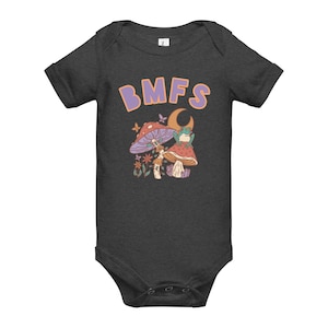 May include: Dark gray baby bodysuit with the text "BMFS" in purple letters. The bodysuit features a colorful illustration of mushrooms, flowers, butterflies, a crescent moon, and a frog. The design has a retro, whimsical style.