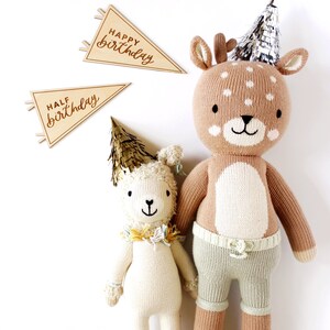 May include: Two knitted stuffed animals wearing party hats. A brown deer with white spots and a white lamb with a gold party hat. The deer is wearing a green and white striped diaper. The animals are holding wooden signs that say "Happy Birthday" and "Half Birthday".