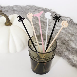 Puede incluir: Un vaso lleno de seis palitos de cóctel con temática de Halloween. Los palitos representan un sombrero de bruja, un fantasma, un murciélago, una calavera, una araña y una calabaza.