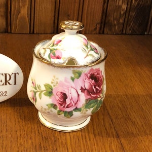 Może przedstawiać: Cukiernica Royal Albert Bone China z pokrywką, zdobiona różowymi i bordowymi różami oraz złotymi obramowaniami. Obok owalny znak z napisem "Royal Albert Bone China England".