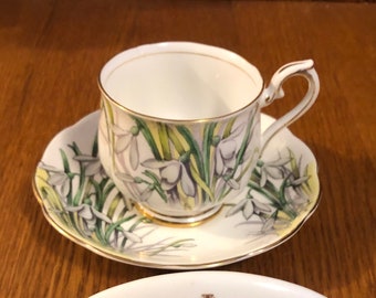 May include: A white teacup and saucer with a floral pattern of white snowdrops on a green background. The saucer has a gold trim. The teacup has a gold handle. The set is made by Royal Albert Bone China, England.