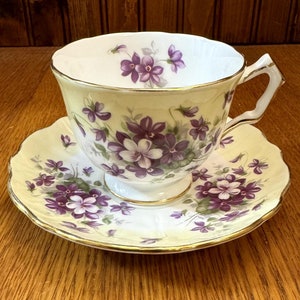May include: A vintage teacup and saucer set. The cup and saucer are decorated with purple floral patterns on a pale yellow background. The edges are trimmed with gold. The cup has a curved handle.