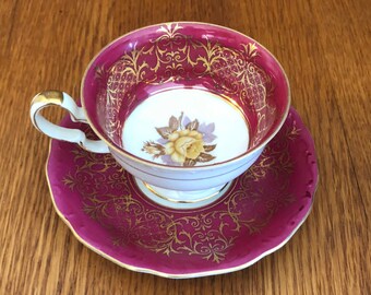 May include: A vintage teacup and saucer set with a pink and gold floral design. The cup and saucer have a white interior and a gold rim.
