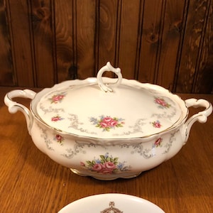 Könnte beinhalten: Weiße Keramik-Servierplatte mit Deckel und Blumenmuster. Die Platte hat zwei Griffe und einen goldenen Rand. Der Deckel hat einen Knopf oben. Die Platte steht auf einer Holzoberfläche. Die Platte trägt ein Etikett mit der Aufschrift "Royal Albert Bone China England".