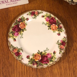 Puede incluir: Un plato de porcelana blanca con un borde dorado y un diseño floral con rosas rojas, rosas y amarillas. El plato está hecho por Royal Albert, Inglaterra.