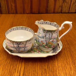 May include: A white porcelain sugar bowl and creamer set with a floral and birch tree design. The set is on a matching tray with a gold trim.