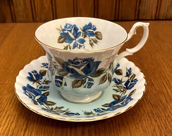 以下が含まれることがあります： ヴィンテージのティーカップとソーサーのセット。白い磁器のカップとソーサーには、金色のトリムが付いた青いバラの花柄が施されています。カップには湾曲したハンドルがあり、お揃いのソーサーの上に置かれています。