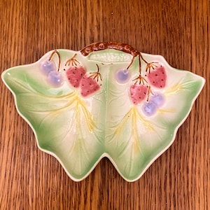 Op de afbeelding: Een groene keramische schaal in de vorm van twee bladeren met een bruine stengelhandvat. De schaal is versierd met roze aardbeien en paarse bosbessen.