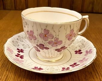 May include: A white teacup and saucer with a floral pattern. The cup and saucer feature a delicate, hand-painted design of purple flowers and grey branches on a white background. The edges of the cup and saucer are trimmed with gold.